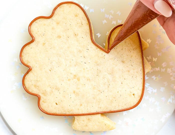 Image of Using the brown detail consistency icing, outline the main body of the turkey shape, as shown in the photo. Steps 3-6 will use a wet-on-wet technique, and will need to be performed immediately following each step, while the icing is still wet. Please read through steps 3-6 before beginning. Pro Tip: With wet-on-wet technique, make sure your flood icing consistencies are all the same to ensure an even, smooth effect.