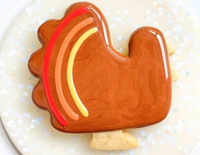 Image of While the brown flood is still wet, pipe 3 curved lines of red, then orange, then yellow flood consistency icing along the left side of the turkey, on its tail, as shown in the photo.