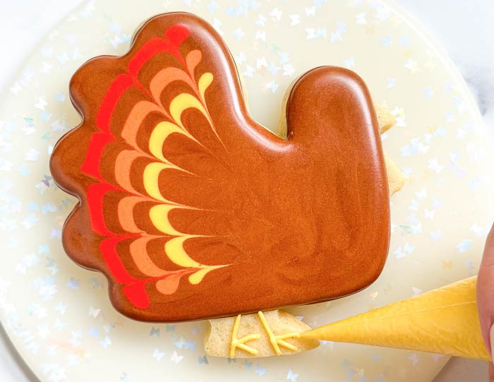 Image of Using the yellow detail consistency icing, pipe the turkey's feet as shown. Feel free to use piping tips or cut your tipless bags to a thickness that is desired. Pipe the turkey's beak and use a scribe tool or toothpick to shape it if needed.