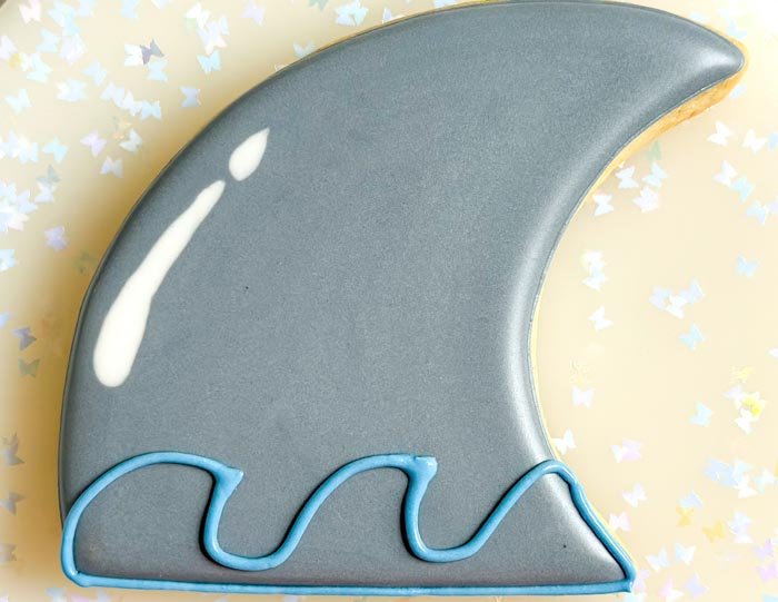 Image of Using blue piping consistency royal icing, pipe a series of ocean waves on the bottom of the fin. These waves do not have to be perfectly piped or symmetrical. Have some fun with it!