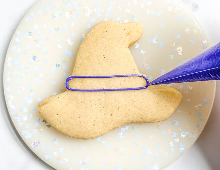 Image of Using the purple detail consistency icing, outline the middle section of the witch hat, as shown in the photo. This will be the hat band.