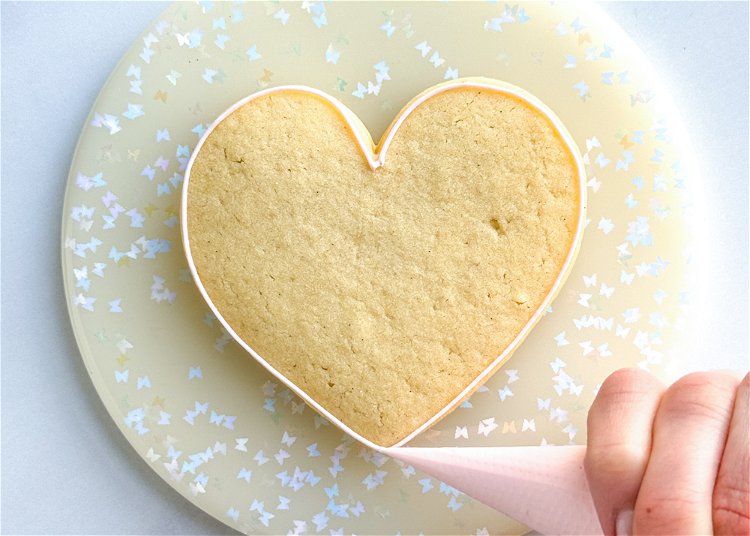 Image of Using the light pink outline consistency icing, outline the heart shape. Hold your piping bag over the heart and let the icing fall down to get clean, precise lines.