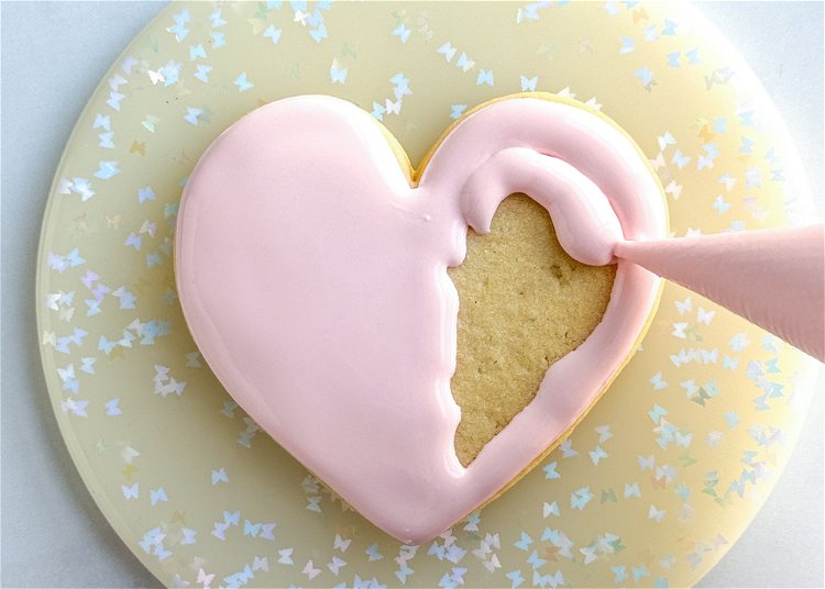Image of Flood the heart shape with the light pink flood consistency icing. Let this flood base dry for 45-60 minutes before moving onto the next step. Drying your cookies in a dehumidifier or under a fan will speed up this process.