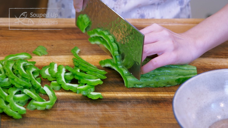 Image of While waiting, we can prepare the bitter melon. Same as before, cut it in half. Remove the white spongy inside part using a spoon. Once it is clean, slice it into thicker strips at a 45-degree angle.