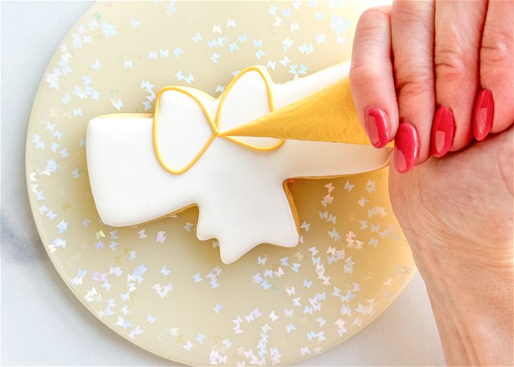 Image of Using the yellow outline consistency, outline the center bow, following the shape of the cookie, as shown in the photo. It may help to hold your piping bag slightly over the cookie, letting the icing fall down onto the cookie. 