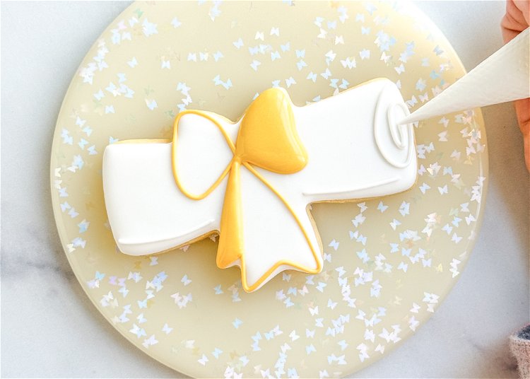 Image of Using the white outline consistency icing, pipe two short lines at the bottom of the design, on either side of the bottom of the yellow bow. Pipe a swirl on the right side of the cookie, as shown in the photo. This detail represents the edges of the diploma paper. 