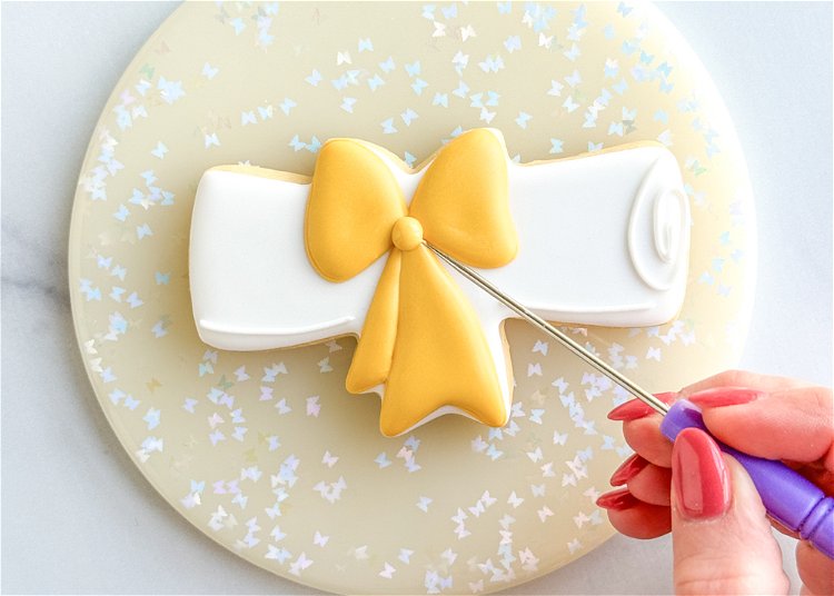 Image of Using the yellow outline consistency icing, pipe a pea-sized dot in the center of the bow to finish the design.  
