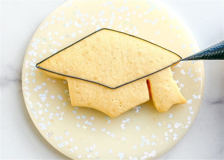 Image of Using the black outline consistency icing, outline the top section of the graduation cap as shown in the photo.