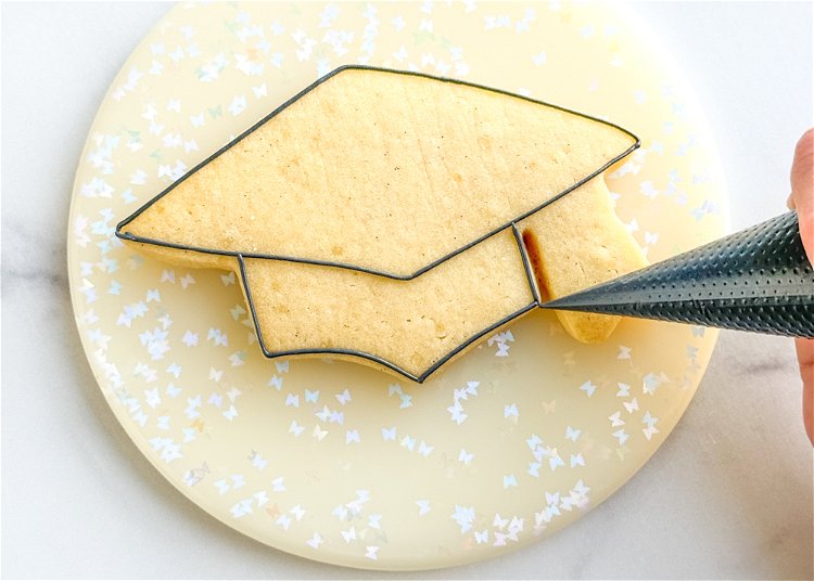 Image of Using the black outline consistency icing, outline the bottom section of the graduation cap. 