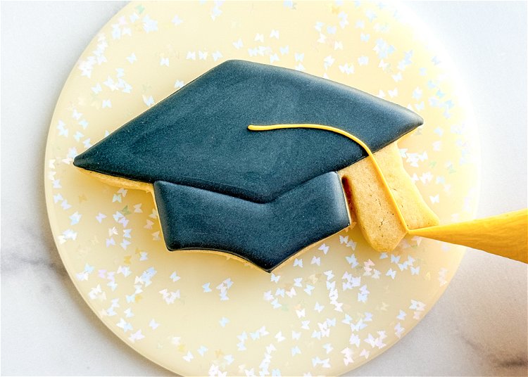 Image of Starting in the middle of the top section of the graduation cap, pipe a line from the center to the bottom right side of the cap with the yellow outline consistency icing.  