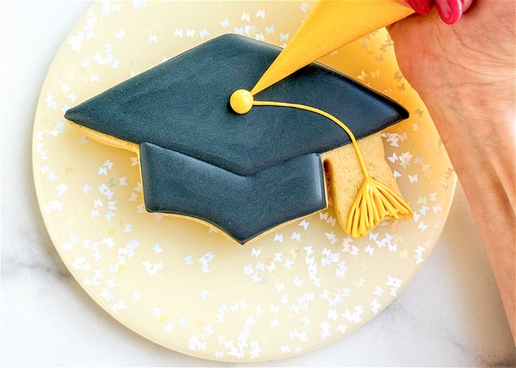 Image of Pipe a small dot of yellow outline consistency icing at the center of the graduation cap, where the tassel line begins. 