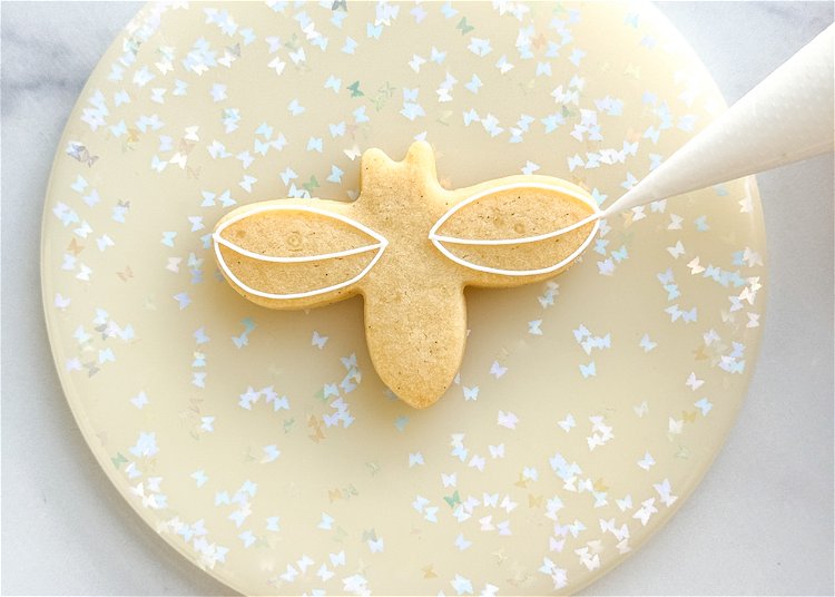Image of Using the white outline consistency icing, outline the wings of the bee as shown in the photo. 
