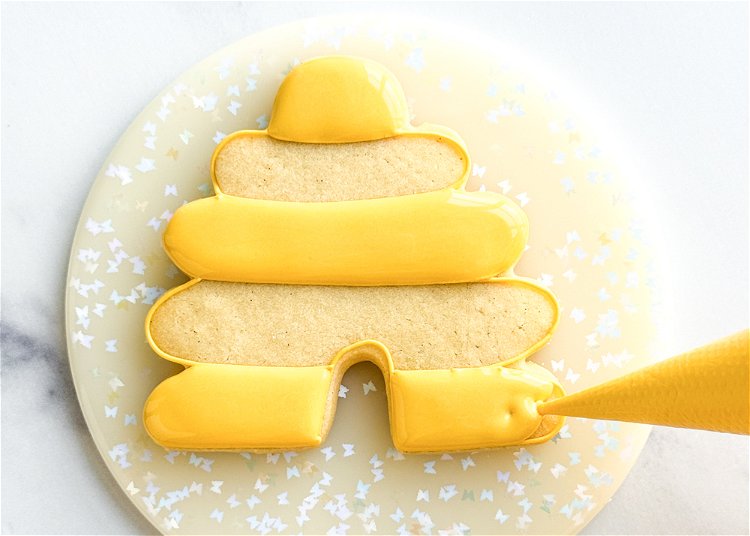 Image of Using the yellow flood icing consistency, fill in the sections of the beehive where you just piped the squiggle of outline consistency icing. Use your scribe tool to settle the icing and pop any air bubbles and to coax the icing to the edges of the cookie. While the flood icing dries, pipe the bee royal icing transfers. Pro Tip: Flooding small sections of a cookie with royal icing leaves the icing vulnerable for craters, which are holes or sinking that can often occur in the icing as it dries. In this design, the elongated oval sections of the beehive are at risk for craters, so adding this squiggle of outline consistency icing helps minimize the risk of craters by providing a cushion for the flood icing so that when it tries to sink as it dries, it has the thicker outline icing underneath to help hold it up. 