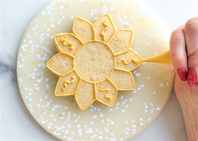 Image of Add a squiggle of yellow outline consistency icing to alternating petals. Pro Tip: Flooding small sections of a cookie with royal icing leaves the icing vulnerable to craters, which are holes or sinking that can often occur in the icing as it dries. In this design, the petal sections are at risk for craters, so adding this squiggle of outline consistency icing helps minimize the risk of craters by providing a cushion for the flood icing so that when it tries to sink as it dries, it has the thicker outline icing underneath to help hold it up. 