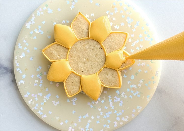 Image of Using the yellow flood consistency icing, fill in the petals you just added the squiggle of outline consistency icing to. Use your scribe tool or toothpick to smooth out the icing of each petal and pop any air bubbles. Allow these sections to crust over for 20-30 minutes before moving onto the next step. If you do not wait between sections, you risk the two wet sections next to each other bleeding into each other, losing definition. 