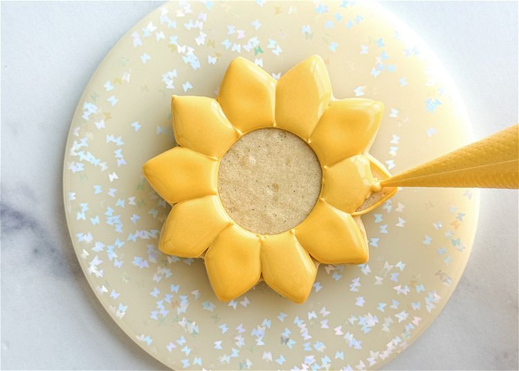 Image of Using the yellow flood consistency icing, fill in the petals you just added the squiggle of icing to. Use your scribe tool or toothpick to smooth out the icing of each petal and pop any air bubbles. Allow these sections to crust over for 20-30 minutes before moving onto the next step. You can speed the drying process by placing the cookies under a fan. 