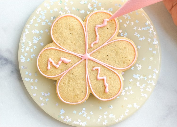 Image of Using the pink outline consistency icing, pipe a squiggle of icing in the center of three alternating petals.  Pro Tip: Flooding small sections of a cookie with royal icing leaves the icing vulnerable to craters, which are holes or sinking that can often occur in the icing as it dries. In this design, the petals are at risk for craters, so adding this squiggle of outline consistency icing helps minimize the risk of craters by providing a cushion for the flood icing so that when it tries to sink as it dries, it has the thicker outline icing underneath to help hold it up. 