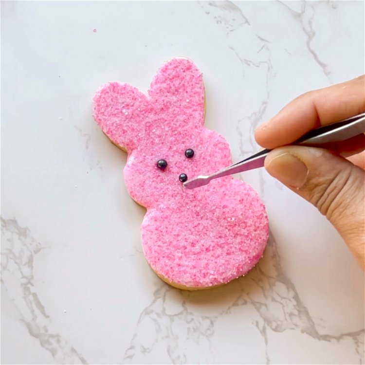 Image of While the icing is still wet, gently place the black pearl sprinkles for the eyes and nose of the bunny. It may help to use tweezers to place them.  Gently push them into place.  Alternatively, you can use black icing for the eyes and nose. Make a divot with a toothpick, and pipe small dots of black icing for the eyes and nose of the bunny. 