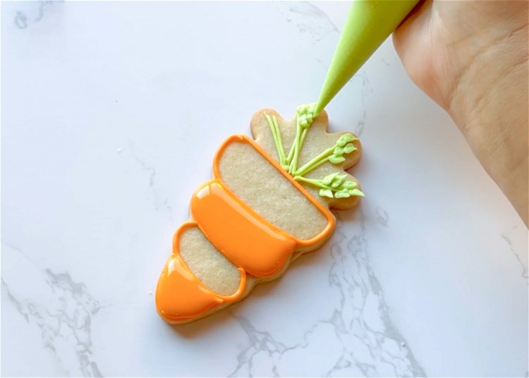 Image of While the two flooded sections are drying, pipe the leaves of the carrot with green outline consistency icing. To create the leaf shape, hold your piping bag and flatten the tip as much as possible. Snip a tiny amount off of each side of the bag to create a small “V” shape. Practice your piping on a paper towel or piece of parchment paper to perfect the shape, applying more or less pressure and adjusting as needed. 