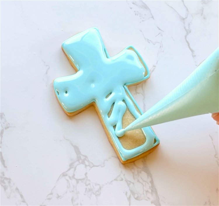 Image of Flood the interior of the cross with blue flood consistency icing. Use a scribe tool or toothpick to gently move the icing around. This ensures full coverage and also pops any air bubbles in the icing.  Wait for this base flood to dry for 30-60 minutes, or until just crusted over. Placing cookies under a fan may help to dry the icing more quickly. 