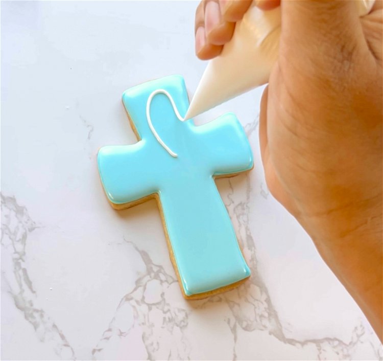 Image of Use your white outline consistency icing to create an elongated figure 8 or infinity symbol shape on the vertical part of the cross.  Start at the center of the cross, and position the piping bag above the cookie so that the icing falls on the cookie, outlining the figure 8 shape. Your white icing should be roughly toothpaste consistency. 