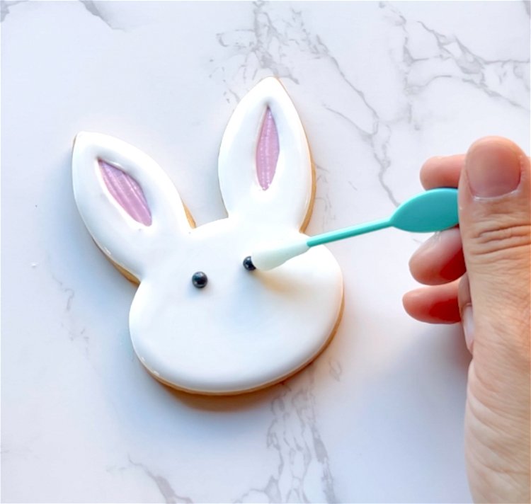 Image of While the icing is still wet, gently place two black pearl sprinkles for the bunny eyes.  Alternatively, you can wait until the base flood is just crusted over, about 30-60 minutes, and pipe two small dots of black outline consistency icing for the eyes.