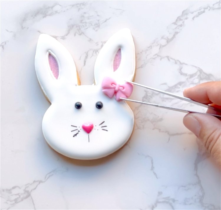 Image of Place your bow royal icing transfer or sprinkle onto the bunny ear. Use white outline-consistency icing to adhere the transfer to the bunny ear. By using the same color as the base (white in this case), you can easily blend and wipe any of the adhesion icing that may get squished out as you are placing your transfer. 