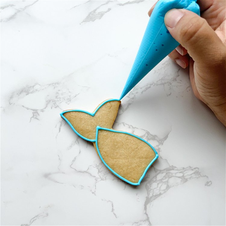 Image of Outline the fins of the mermaid tail with teal outline consistency icing. Once complete, you should have two distinct areas outlined with teal icing. 