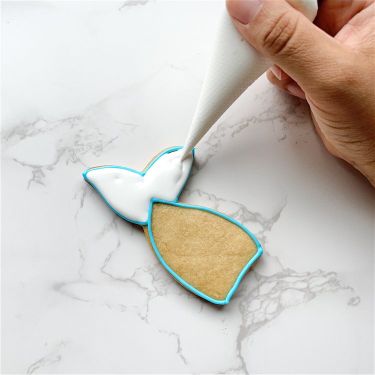 Image of Using white flood consistency icing, flood the fins of the mermaid tail. Use a scribe tool or toothpick to gently move the icing around, coaxing it to the edges and popping any air bubbles. 