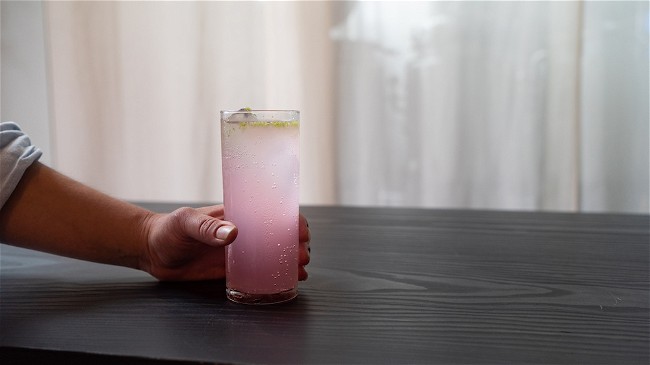A Collins glass with a lovely purple color, the vibe is moody. A hand holds the glass but the person is out of sight.