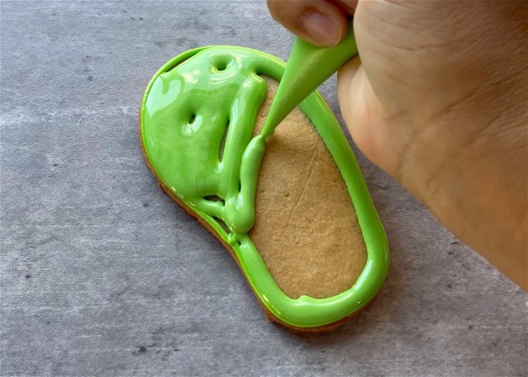 Image of Flood the shape with green royal icing, using a scribe tool or toothpick to move the icing around, ensuring full coverage and popping any air bubbles. Move quickly to the next steps through Step 7, as the icing needs to be wet for this design to work.  