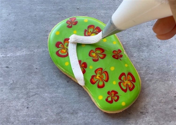 Image of Once the icing has dried to the point of just crusting over, you can pipe the sandal straps. Use a piping bag fitted with a small star tip to get the correct texture. Pipe two lines of white icing that meet in the upper part of the sandal to mimic a flip flop sandal.  