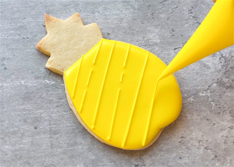 Image of Use your yellow outline consistency icing to create diagonal lines along the pineapple shape. Pro tip: To create straight, even lines hold your piping bag above the shape and let the icing fall into place, rather than holding the piping bag close to the cookie. 