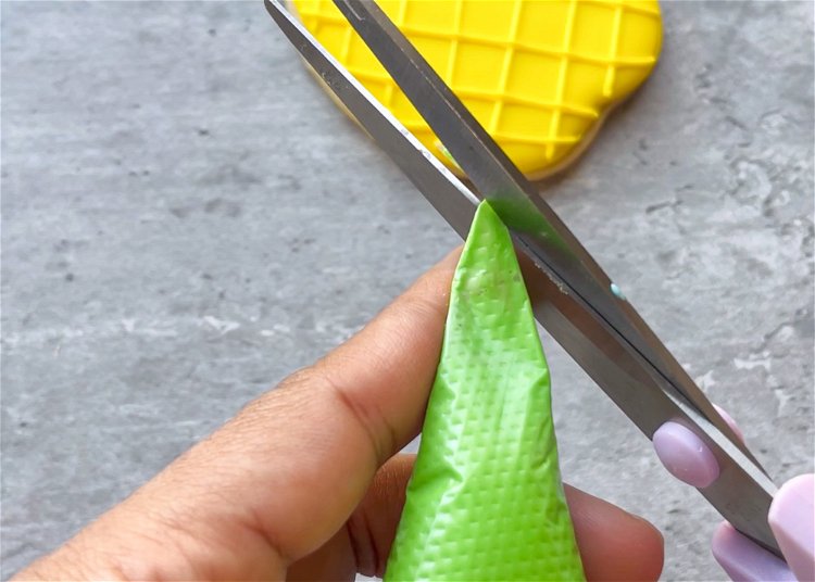 Image of Snip a “V” shaped tip off of the end of your green royal icing tipless piping bag. This shape creates a ruffled look that is great for leaves. Pro tip: Snip a little at a time, as you can always remove more of the tip, but you can’t add more back on. It may help to do some test runs on a paper towel or piece of parchment paper to ensure you’re getting the correct texture and width for your leaves. 