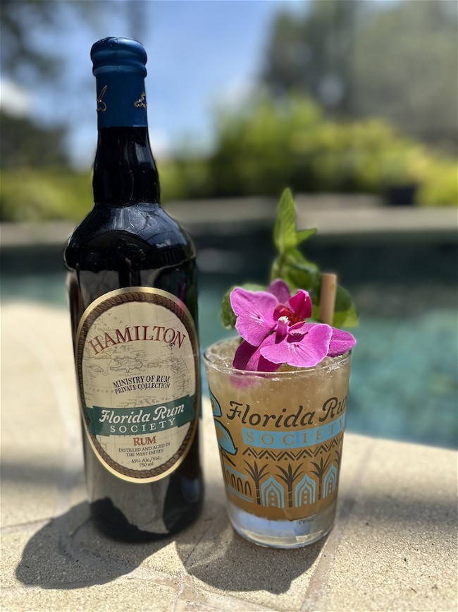 A tiki looking cocktail next to a bottle of Hamilton Florida Rum Society rum, a pool in the background