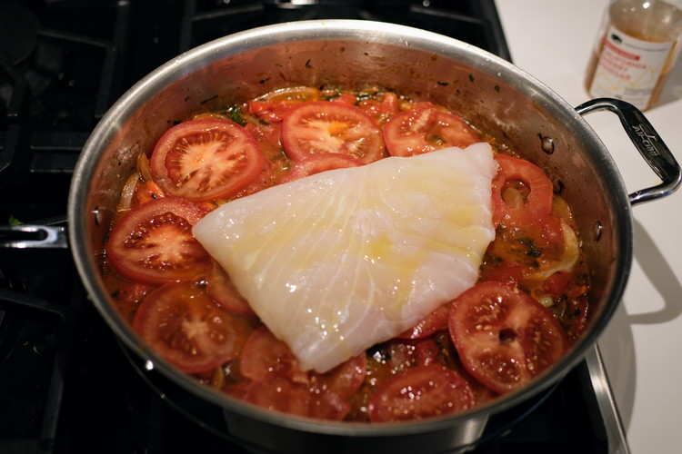 Image of Shut off stove top. Carefully spread out the sliced tomatoes over your cooked sauce so they create one layer.  Lightly salt the tomatoes.  Place the cod loin over the sliced tomatoes and lightly drizzle some olive oil over the fish.