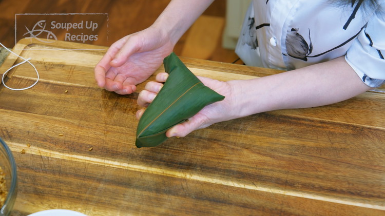 Image of Fold the upper leaves down and stay there with some force to establish the shape. The hand at the bottom should squeeze the zong zi. Use the hand on the top to carefully fold the edge done. Do the same...