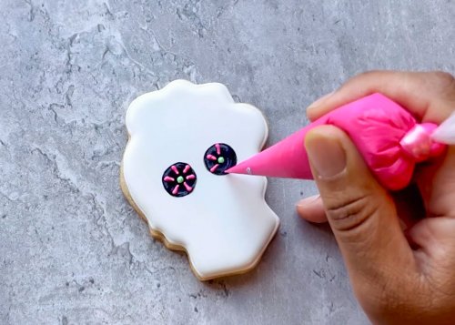 Image of Use your pink royal icing to pipe four lines extending outward from the green dot in the center of each eye hole. Pro tip: To get precision lines, start by cutting off a very small amount of your tipless piping bag. Do a test pipe on a paper towel or piece of parchment paper. If the line is squiggly as you pipe, the hole is too small and will need to be cut larger until you can pipe a consistent straight line. When piping lines, hold the piping bag over the cookie and let the icing “fall” onto the cookie. This creates evenly thick lines. You can use a scribe tool to gently coax any wayward icing into place. 