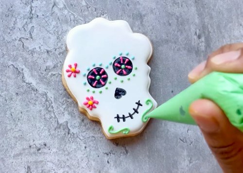 Image of Add flowers to the cheeks of the skull using your pink and yellow royal icing. Pipe lines around the mouth using your green royal icing. Use the same technique mentioned in step 5—let the icing fall into place, and coax any drips or mistakes into place using a scribe tool before the icing dries. 