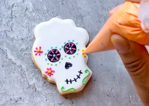 Image of Begin decorating the flower crown by piping a small yellow flower on the right side of the sugar skull.  