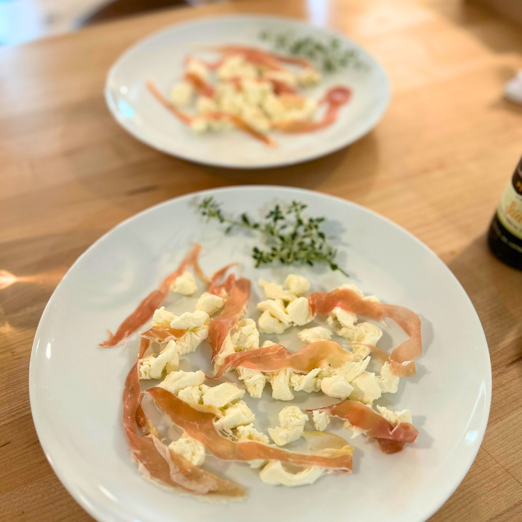 Image of To serve; spoon cheese (or place slices of fresh mozzarella) onto a plate, layered with strips of Prosciutto di Parma, then place on top the potatoes, olives, tomato and cod and garnish with crispy capers and black garlic sauce.