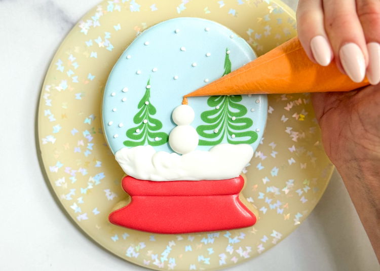 Image of Using the orange outline consistency icing, pipe a small squiggle on top of the second white dot you piped. This is meant to be the snowman’s carrot nose. See, our snowman is looking up at the falling snow! 