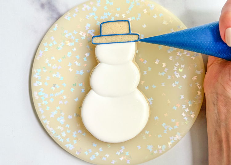 Image of Using the royal blue outline consistency icing, outline the snowman’s hat in two sections, as shown in the photo. Add a squiggle of outline consistency icing in the top section of the hat.  Pro Tip: It is best practice to add a squiggle of outline consistency icing before flooding small areas of a cookie to help support the flood icing and prevent cratering, which are holes or sinking that can occur when flood icing dries. Cratering is much more likely to occur when flooding a small area of a cookie, so that is why we use this trick to help prevent it in these small sections. 