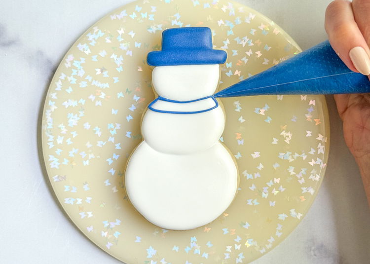 Image of Using the royal blue outline consistency icing, pipe a scarf around the snowman’s neck, as shown in the photo. Add a squiggle of icing in the middle of the section. 