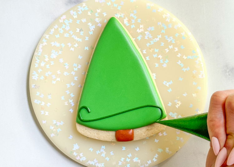 Image of Using the dark green outline consistency icing, pipe a swirling line in a continuous motion, moving from the bottom of the tree to the top. Pro Tip: You may find you need to practice this on a piece of paper towel before you attempt this design on the actual cookie. Make sure you’re comfortable with the size of the hole in your piping bag, as well as the consistency of your icing. Your icing should be a consistency similar to toothpaste. Learn more about royal icing consistencies here.   If the icing is difficult to control, it may be too thin. Add powdered sugar, a little bit at a time, to achieve the proper consistency.  If your icing is difficult to push out of the bag or comes out in squiggles, it may be too thick (add water a little bit at a time to thin out), or the hole in your piping bag may be too small.  