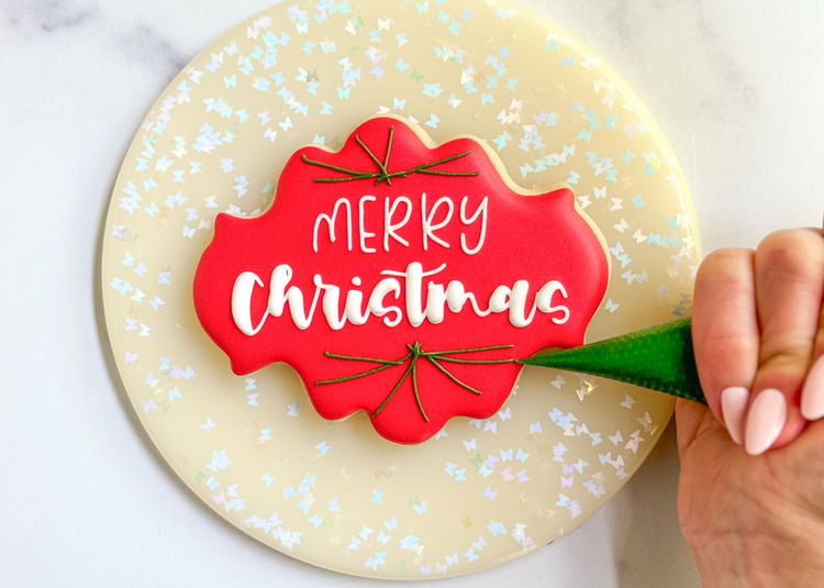 Image of We will finish this cookie design by piping some holiday greenery above and below our lettering. Using the dark green outline consistency icing, pipe some lines for the shape of your greenery. 