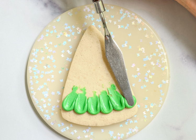 Image of Immediately after piping, use your palette knife or other tool to gently press down on each dot and drag it up and towards the center point of the triangle. Wipe any excess icing off your palette knife or other tool, so leftover icing doesn’t dry on it or affect our design.  