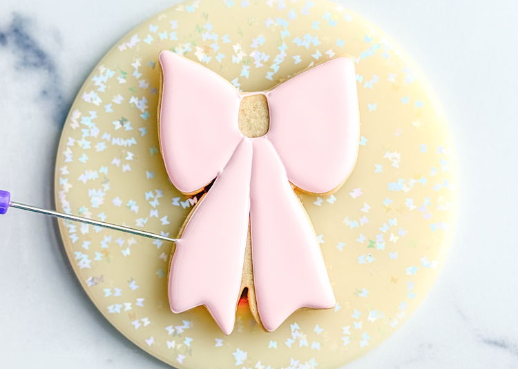 Image of Using the light pink flood icing, fill in the bottom left section of the bow. Use your scribe tool or a toothpick to smooth out the icing and pop any air bubbles that come up. Allow this section to dry for 20-30 minutes before moving onto the next step. 