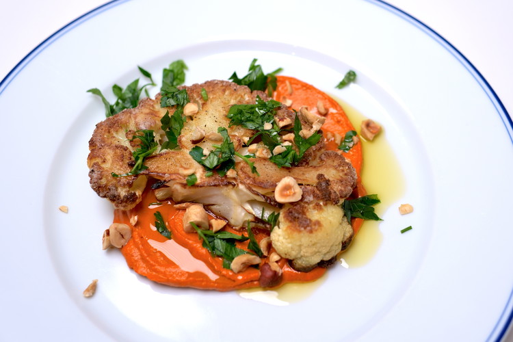 Image of To plate, spread a few large spoonfuls of Romesco sauce on a plate. Carefully put the cauliflower steak on top. Spread chopped hazelnuts and chopped parsley on top.  Drizzle a small amount of olive oil over. Serve
