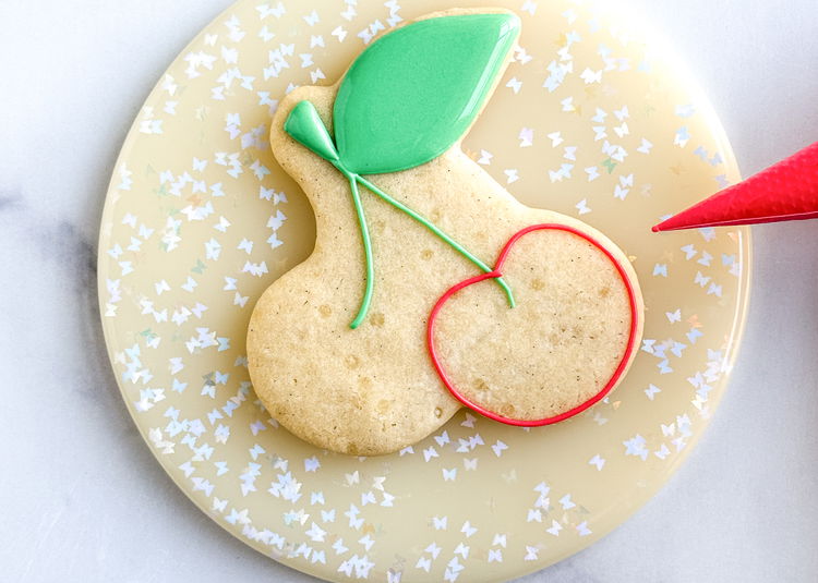 Image of Using the red outline consistency icing, outline the section for the two “side-by-side” cherry sections.  Use the shape of the cookie as the guide for piping these sections. 